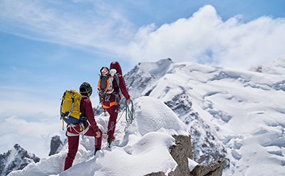 Um grupo de montanhistas com equipamento colorido sobe a um pico montanhoso e nevado sobre um fundo de céus cinzentos.