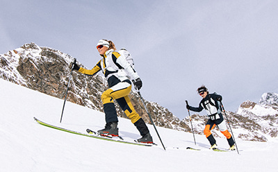 Dois esquiadores com equipamento de inverno colorido estão a esquiar por uma encosta montanhosa sobre um fundo de picos robustos cobertos de neve.