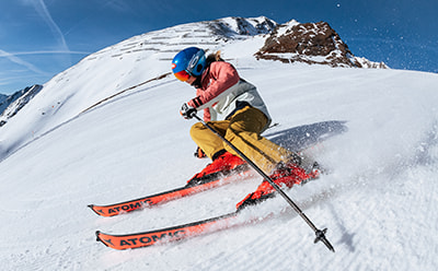 Uma esquiadora com equipamento colorido a esculpir a neve profunda numa encosta montanhosa com picos nevados ao fundo.
