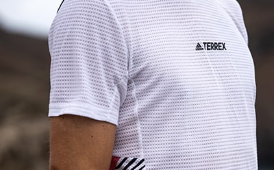Ein weißes, gemustertes Hemd mit dem adidas TERREX Logo ist vor einem verschwommenen Hintergrund im Vordergrund zu sehen.