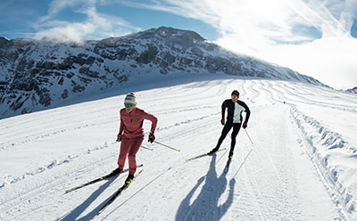 Två skidåkare på snöiga bergsleder, med snötäckta toppar i bakgrunden under en ljus, molnig himmel.