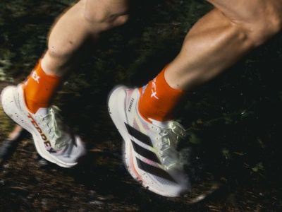 The image shows a person's lower body wearing white and orange athletic shoes, standing on a dirt path surrounded by greenery.