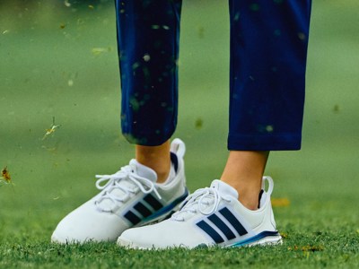 The image shows a person's legs and feet wearing white sneakers with blue stripes, standing on a grassy field with a blurred, green background.