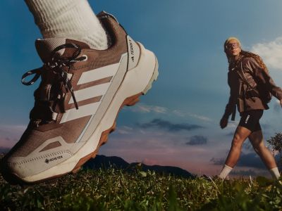 A large hiking boot stands in the foreground, with a person walking in the background against a scenic mountain landscape.