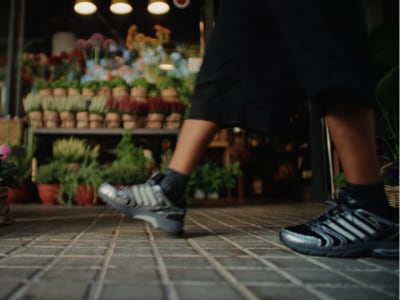 The image shows a person's legs and feet wearing sneakers standing in front of a display of various potted plants and flowers in a store or market setting.