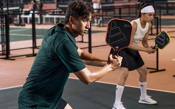 Two individuals, a man and a woman, are engaged in a pickleball match on an outdoor tennis court. The man is wearing a green shirt and the woman is wearing a white shirt, and they both appear to be focused on the game.