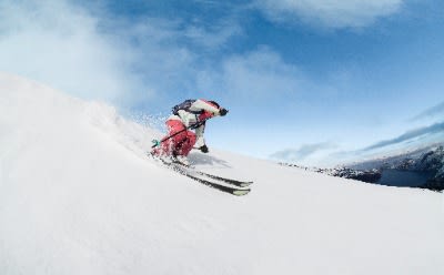 Una persona con chaqueta roja esquiando por una pendiente nevada con un fondo de cielo azul claro con nubes nevadas.