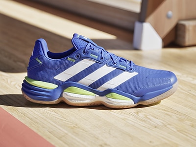 A pair of blue and white Adidas trainers with lime green accents sits on a wooden floor, with a blurred background of what appears to be a room or hallway.