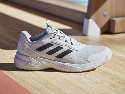 A pair of white and black Adidas tennis shoes rests on a wooden floor, with a blurred background suggesting an indoor setting.
