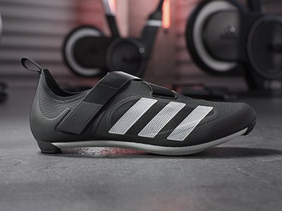 A pair of black and white athletic shoes with a textured sole sits on a concrete floor, with weight training equipment visible in the background.