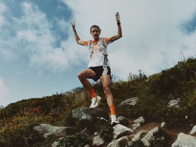 A person in athletic clothing stands triumphantly on a rocky outcrop, with a scenic mountainous landscape visible in the background.