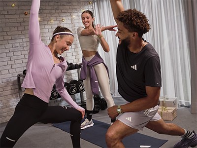 The image shows three people engaged in a fitness activity, with one person in a pink outfit and the other two in athletic wear, in a room with a brick wall and exercise equipment visible in the background.