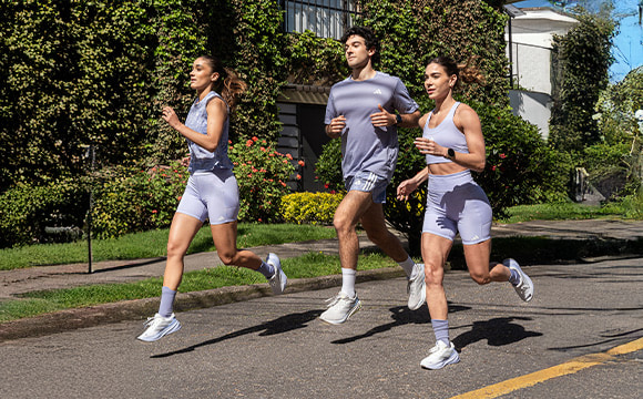 Three young women in athletic wear are jogging together on a paved path surrounded by lush greenery and a building in the background.