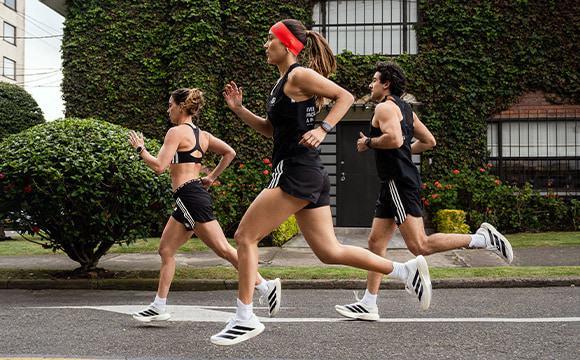 Three young women in athletic attire are jogging together on a paved path surrounded by lush greenery and buildings in the background.