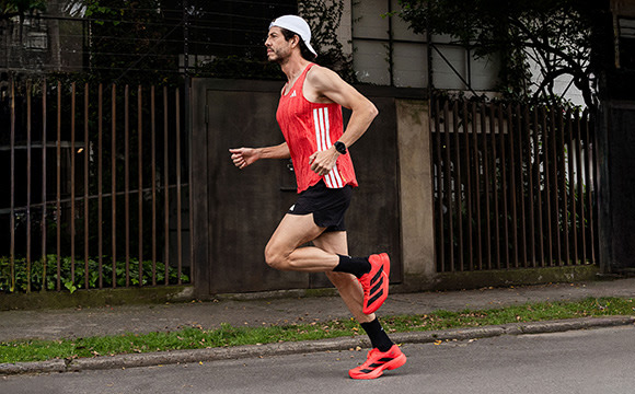 A person in a red and white athletic jersey and shorts is running on a sidewalk next to a metal fence, with trees and buildings visible in the background.