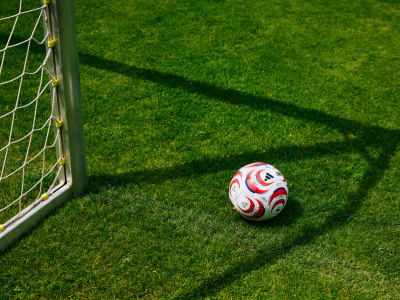 A soccer ball rests on a lush, green grass field, with a soccer goal post visible in the background.