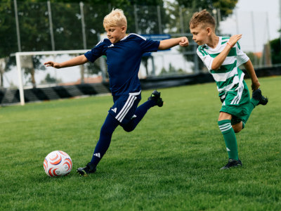 Two young boys in sports uniforms are playing soccer on a grassy field, with a soccer ball in the foreground.