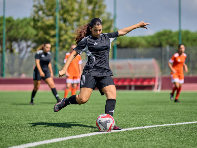 A female soccer player in a black uniform is dribbling the ball on a green artificial turf field, with other players in the background.