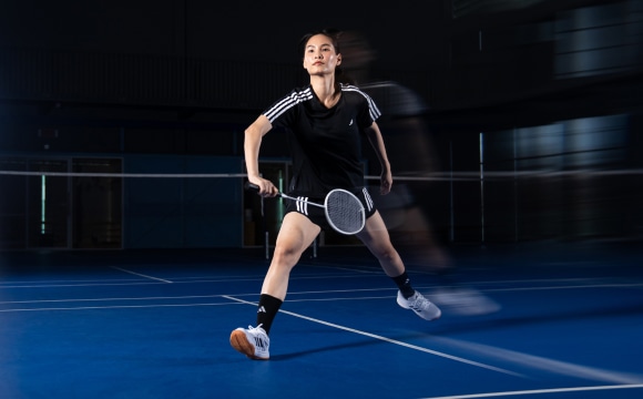 A female athlete in athletic attire is standing on a badminton court, holding a badminton racket and preparing to make a shot.