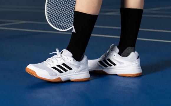 The image shows a person's feet wearing white and black athletic shoes on a blue tennis court, with a tennis racket visible in the background.