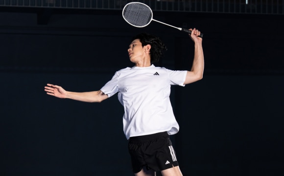 A person in a white shirt and black shorts is holding a badminton racket and appears to be in the middle of a swing on a dark background.