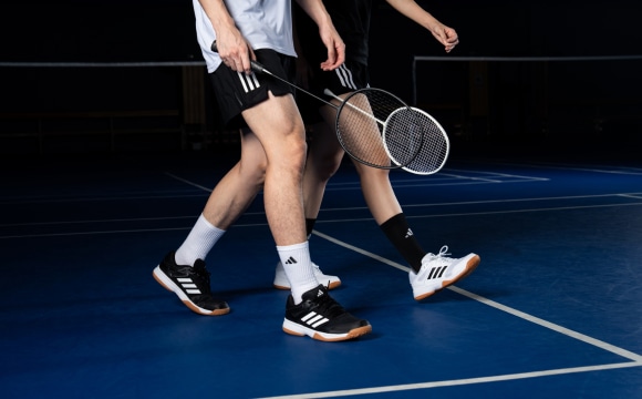 The image shows the lower body of a person wearing athletic clothing and holding tennis rackets on a blue tennis court.