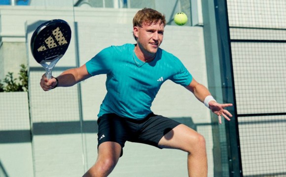A young man in a teal shirt is playing paddle tennis on an outdoor court with a glass wall in the background.