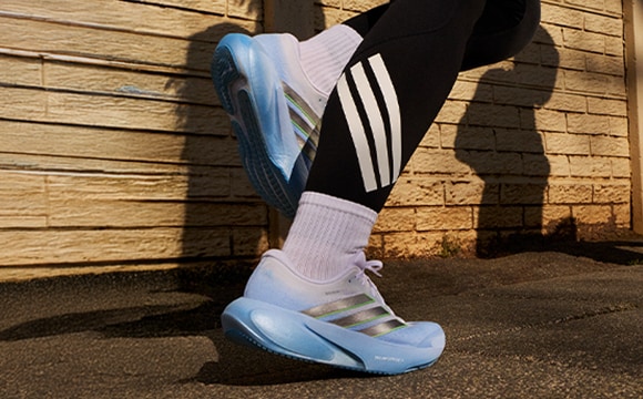A pair of colorful athletic shoes with distinctive stripes and patterns stands against a wooden wall, casting a long shadow on the ground.