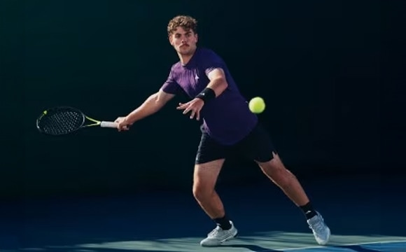 A tennis player in a purple shirt and shorts stands on a dark court, preparing to hit a tennis ball.