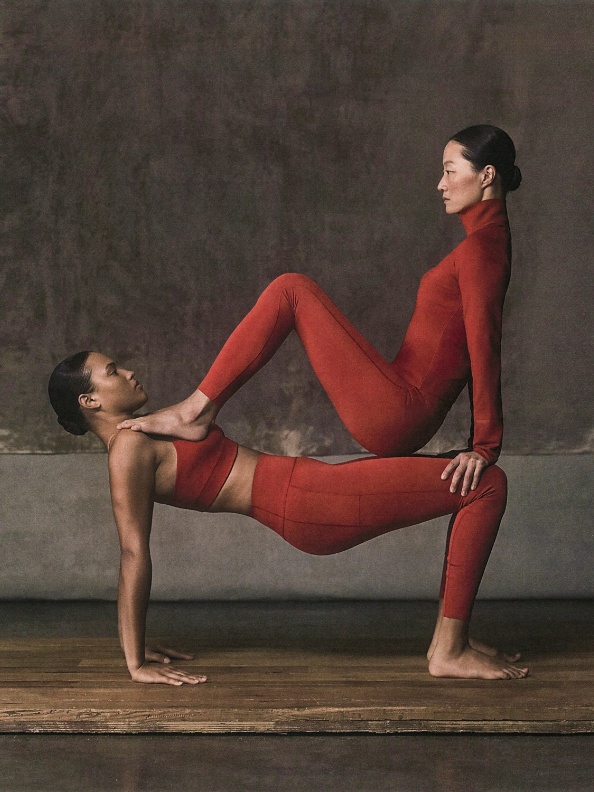The image depicts two individuals in red bodysuits performing a yoga or acrobatic pose on a wooden floor against a dark background.