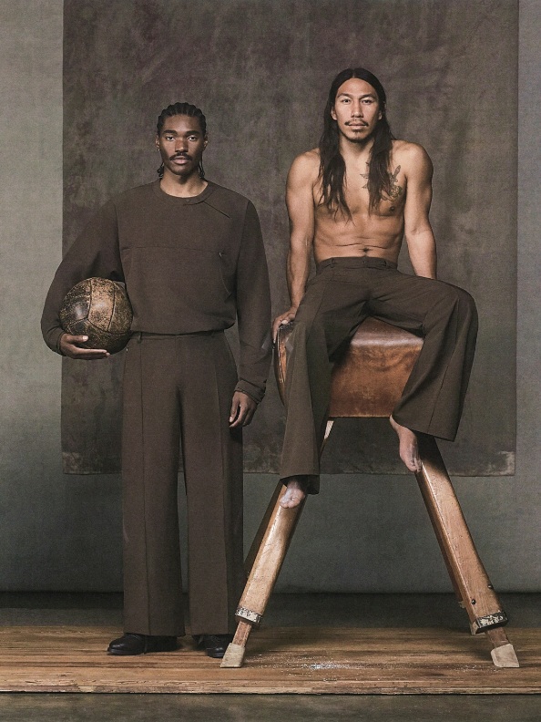 Two individuals, one wearing a dark outfit and the other in a more casual attire, are standing on a wooden platform against a plain background.