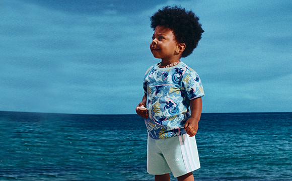 A young child with curly hair stands on a beach, wearing a colorful patterned shirt, against the backdrop of a vast, serene ocean.
