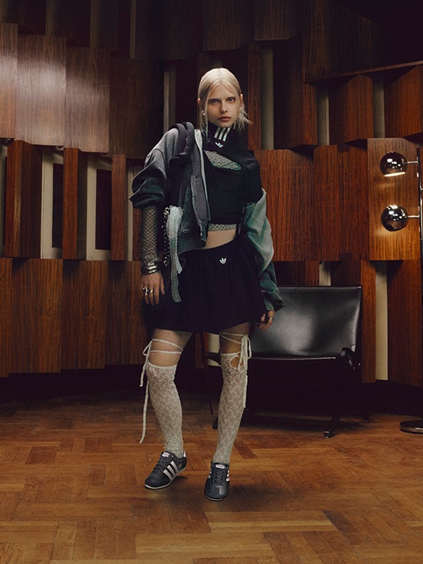 A person wearing a green jacket, black pants, and white sneakers stands in a room with wooden paneling and furniture.