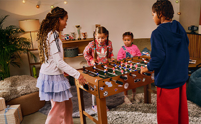 A group of children, two girls and two boys, are gathered around a foosball table, engaged in a lively game, with a cozy living room setting in the background.