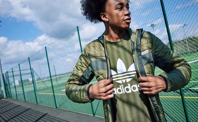 A person wearing an Adidas jacket stands in front of a fenced sports court against a backdrop of a cloudy sky.