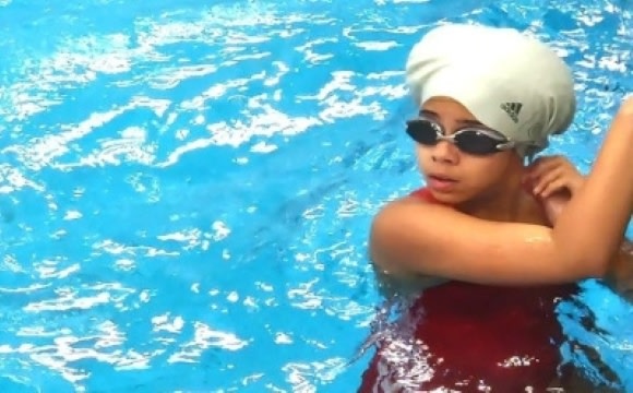 A person wearing a red swimsuit and sunglasses stands in a pool with clear blue water and a bright sky in the background.