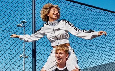The image shows a person with curly hair wearing a gray athletic jacket, jumping and smiling in front of a blue wire mesh fence.