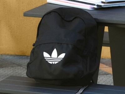 Incluye una mochila negra con el logotipo de adidas sobre un escritorio o un estante, con libros y otros objetos visibles al fondo.