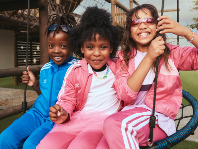Tres niñas pequeñas, todas con ropa brillante y colorida, sonríen y posan juntas en lo que parece ser un patio de recreo o un entorno al aire libre con una valla o una estructura visibles en el fondo.