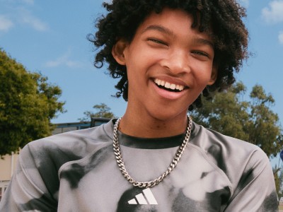Un joven sonriente con el pelo rizado, vestido con una camisa gris y un collar, parado frente a los árboles y un cielo azul.