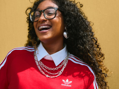 Una persona sonriente con el pelo rizado y gafas con una chaqueta roja de adidas se coloca de pie sobre un fondo amarillo.