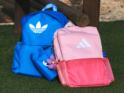 Two colorful Adidas backpacks, one blue and one pink, are placed on a grassy surface in front of a wooden structure.