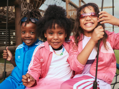 Three young girls, each with distinct hairstyles and wearing colorful apparel, are smiling and posing together in what appears to be an outdoor setting with a wooden structure in the background.