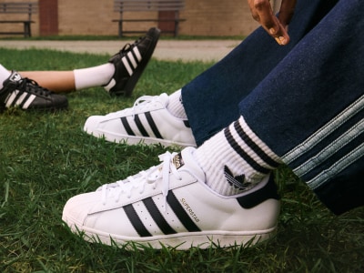 L'image montre une paire de sneakers Adidas blanches avec des bandes noires sur un terrain en herbe, avec les jambes et les pieds d'une personne visibles au premier plan.