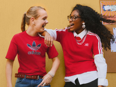 Two young women wearing red Adidas shirts are standing together and smiling in what appears to be a classroom or school setting, with colorful posters and artwork visible in the background.