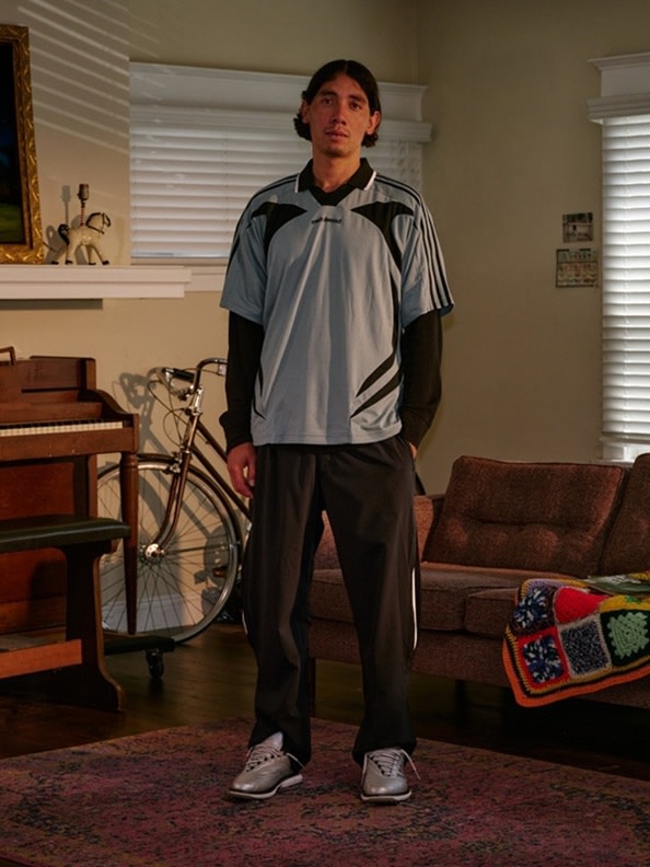 A young man wearing a gray and black athletic jacket stands in a room with a bicycle and various other objects in the background.
