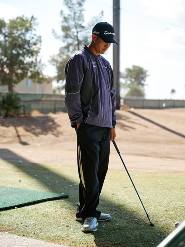 A person wearing a dark-colored jacket and cap is standing on a golf course, with trees and buildings visible in the background.
