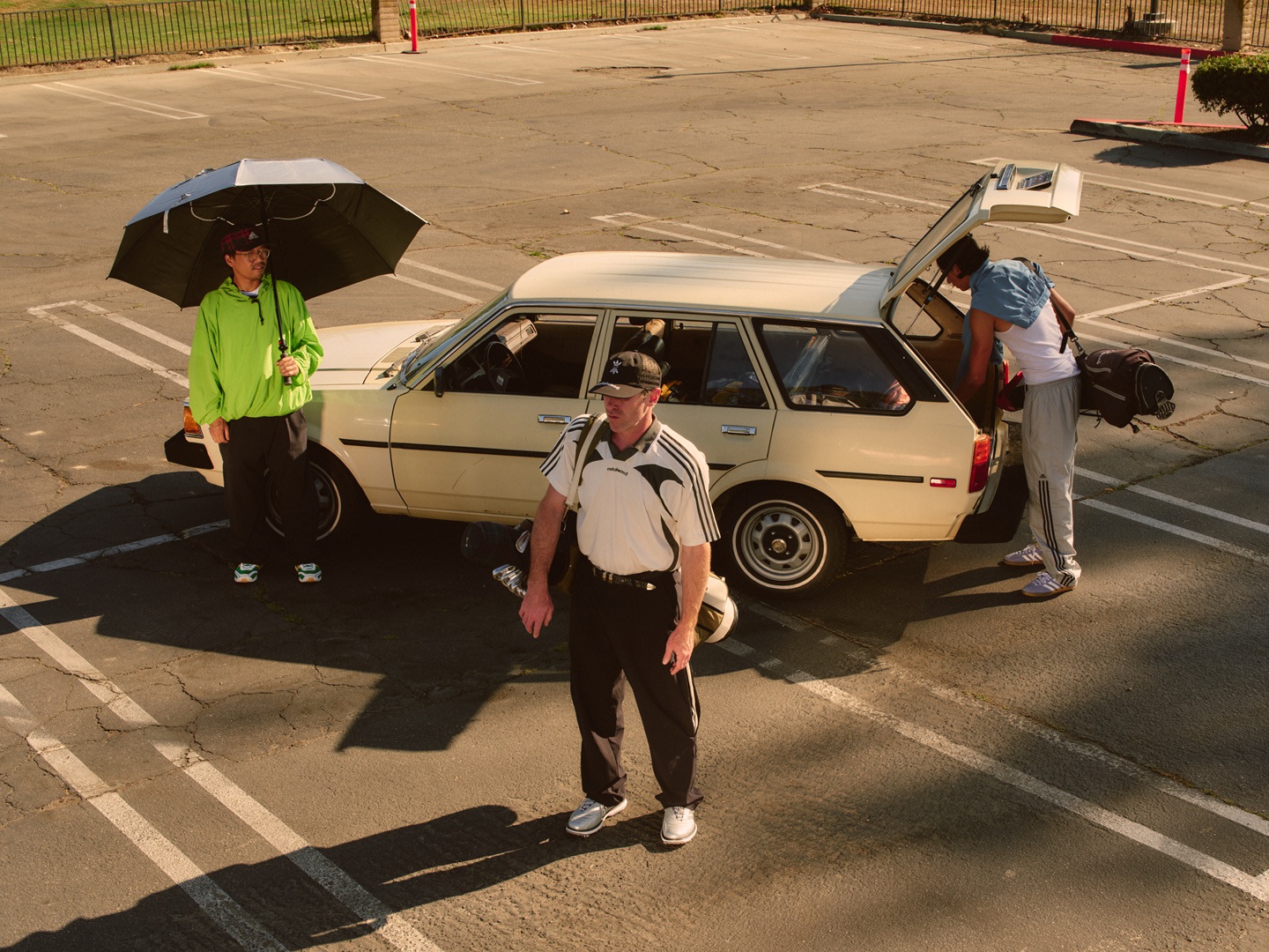 The image shows a group of people standing around a broken-down car in a parking lot, with one person holding an umbrella to shield themselves from the sun.