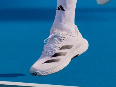 Une sneaker adidas blanche avec des bandes noires est suspendue sur un fond bleu vif.