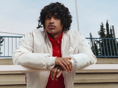 La imagen muestra a un joven de pelo oscuro rizado con un abrigo blanco y una corbata roja, sentado en unas escaleras con una barandilla de fondo.